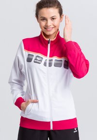 Weiße Sportjacke mit pinken Akzenten, die einen hohen Kragen, einen Reißverschluss und vertikale Streifen aufweist. Hat seitliche Taschen und ein Logo auf der Brust.