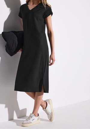 Czarna dzianinowa sukienka z dekoltem w kształcie litery V i krótkimi rękawami, z bocznym rozcięciem. Połączona z białymi i beżowymi sneakersami. Teksturowane wykończenie tkaniny.