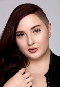 Porträtt av en person med långt brunt hår och en rakad sida, iförd en svart texturerad topp med metallaccenter, lätt smink och en läpppiercing.