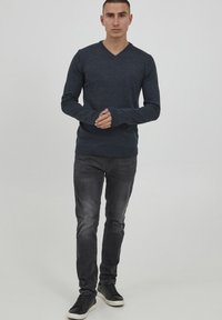Maglione grigio a scollo a V realizzato in tessuto morbido, abbinato a jeans neri sbiaditi e sneakers scure. Maniche lunghe con polsini a coste, design semplice.