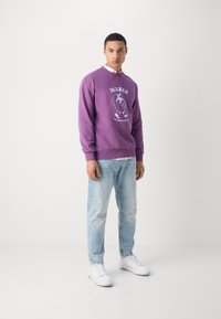 Lilla hettegenser med grafisk design foran, kombinert med lyseblå slitte jeans og hvite joggesko. Stilen er avslappet og moderne.