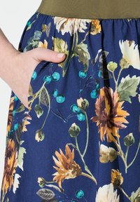 Kukallinen hame tummansinisellä pohjalla, jossa on vihreitä, keltaisia ja valkoisia kukkia. Sisältää sivutaskut ja tiukasti istuvan oliivinvihreän vyötärön.