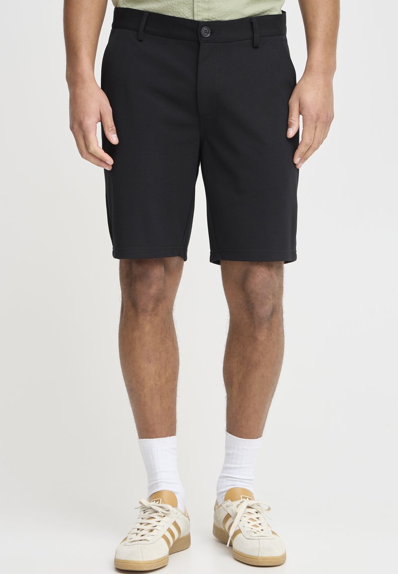 Shorts noirs en tissu doux, avec un devant plat, des passants de ceinture et une fermeture à un bouton. Portés avec des baskets beiges et des chaussettes blanches.