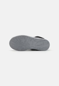 Zapato negro con un acabado texturizado en la parte superior y una suela de goma gris que presenta un diseño de tracción con patrón de estrellas. La suela es plana y ligeramente redondeada en la punta.