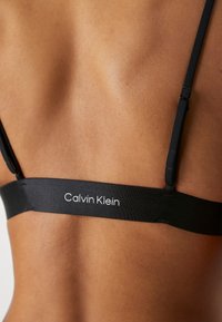 Zwarte bh-band met "Calvin Klein" merknaam, voorzien van verstelbare banden en een gladde textuur tegen gebruinde huid.