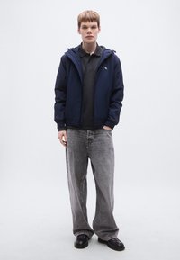 Ναυτικό μπλέ αδιάβροχο με κουκούλα, κλείσιμο με φερμουάρ και ελαστικές μανσέτες. Φορεμένο πάνω από γκρι πόλο, συνδυασμένο με ανοιχτό γκρι τζιν με φαρδιές παντελόνες και μαύρα παπούτσια.