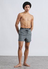 Világosszürke fürdőnadrág könnyű anyagból; fekete rugalmas derékpánttal és zsinórral; sima tapintású, térd fölötti hosszúságú.