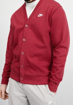 Uomo con cardigan rosso Nike abbottonato sopra una camicia bianca, abbinato a pantaloni della tuta Nike grigio chiaro, con una mano in tasca, su sfondo neutro.