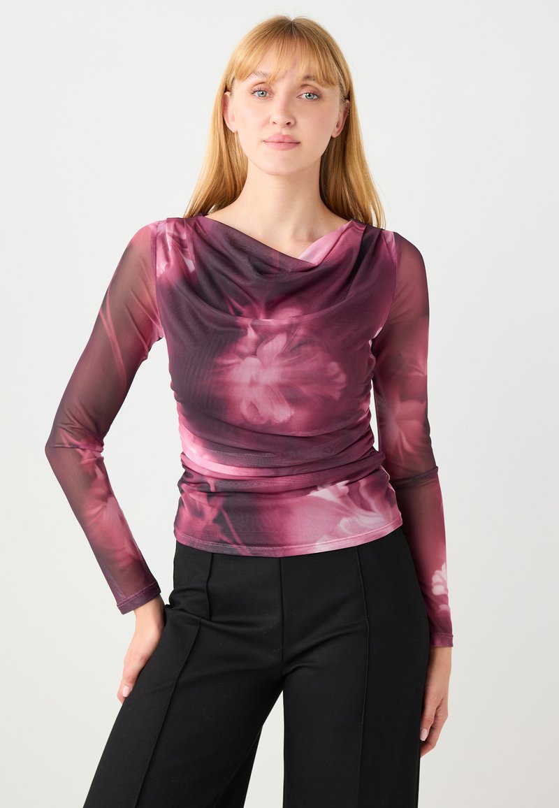 Haut transparent à manches longues dans des nuances de rose et de violet avec un motif floral, col drapé et détails froncés. Associé à un pantalon noir.