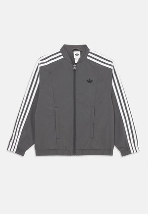 Grijze Adidas trainingsjack met witte drie-strepen op de mouwen, rits aan de voorkant, elastische manchetten en het Adidas-logo op de linkerborst.