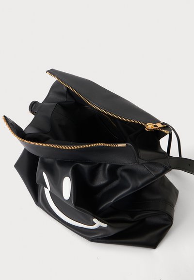 Sac en cuir noir avec un motif visage souriant blanc, fermeture éclair dorée ouverte et intérieur noir visible sur un fond clair uni.
