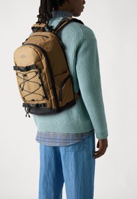 Kamerkleurige rugzak met verstelbare schouderbanden, zwarte accenten, bungee koorddetails en meerdere zakken; gedragen over een gestructureerde teal trui en een gelaagd shirt.