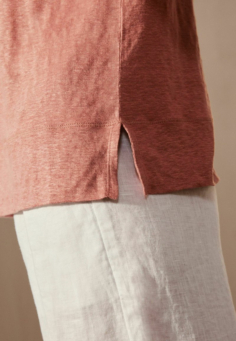 Vue de profil d'une personne portant une chemise couleur rouille avec une petite fente latérale, associée à un pantalon beige clair.