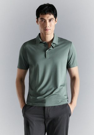 Hombre con una camiseta polo de manga corta verde oliva y pantalones gris oscuro de pie frente a un fondo liso gris claro.