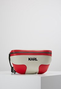 Riñonera hecha de suave cuero sintético, con un cuerpo crema y llamativos acentos rojos, y una cremallera negra, marcada con "KARL" en negro.