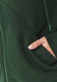 Sudadera con cremallera de color verde oscuro con un bolsillo, que presenta una textura suave, capucha ajustable y costuras de contraste a lo largo de las costuras.