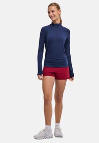 Granatowa sportowa koszulka z długim rękawem i suwakiem do ćwierć długości, w połączeniu z czerwonymi szortami. Modelka nosi białe buty do biegania. Gładka faktura, dopasowany krój.
