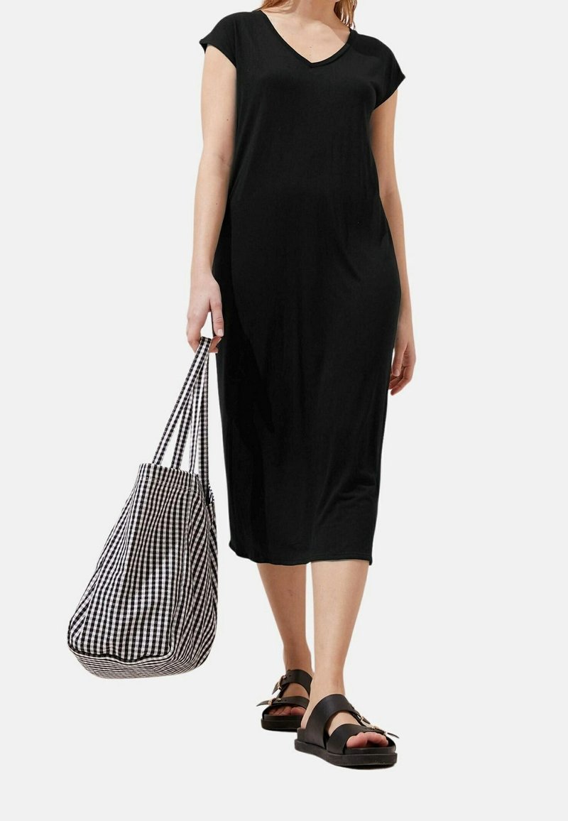 Femme portant une robe midi noire à manches courtes, des sandales noires et portant un grand sac cabas à carreaux noirs et blancs.