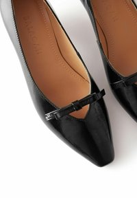 Ballerine nere in pelle verniciata con punta a forma affusolata, dotate di un dettaglio intagliato e un piccolo fiocco decorativo. Interno liscio con rivestimento beige.