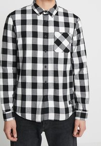 Camicia a maniche lunghe bianco e nero a scacchi realizzata in tessuto, con chiusura a bottoni sul davanti e taschino sul petto sinistro con un motivo a scacchi più piccolo.