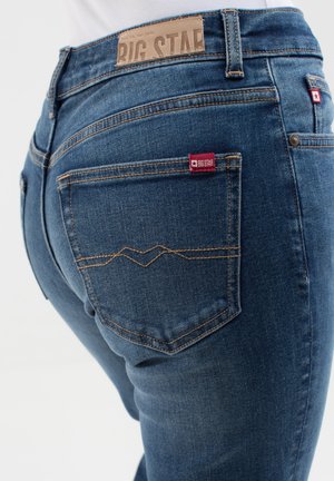 Jeans in denim blu scuro con una texture liscia, caratterizzati da un'etichetta in pelle in vita e da etichette rosse sui taschini posteriori.