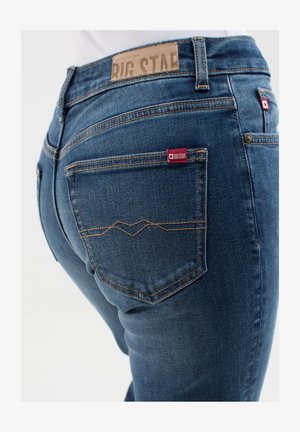 Dark blue denim jeans with a smooth texture, featuring a leather label at the waistband and red logo tags on the back pockets.