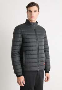 Zwarte gewatteerde jas met hoge kraag, ritssluiting, zijzakken en geribbelde manchetten. Bevat een horizontaal doorgestikt patroon. Rood logo accent.