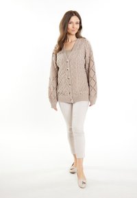 Cardigan beige lavorato a maglia con motivo a trecce, apertura con bottoni, scollatura a V e maniche lunghe. Abbinato a pantaloni aderenti chiari e scarpe.