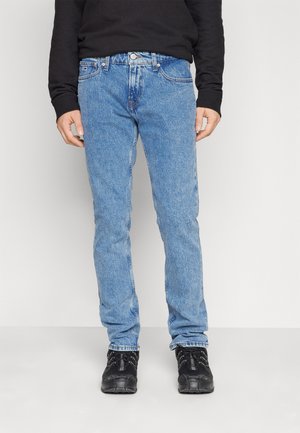 Personne portant un jean bleu clair, un haut à manches longues noir et des baskets noires, debout devant un fond clair uni.