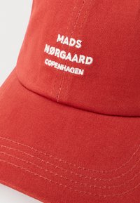 Red cotton cap with a curved brim. Features white embroidered text "MADS NØRGAARD COPENHAGEN" on the front. Textured fabric with visible stitching.