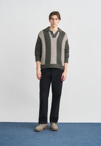 Texturált, olíva zöld és bézs csíkos pulóver V-nyakkivágással és gallérral, fekete nadrággal és világosszürke cipővel, kék szőnyegen.