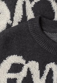 Gros plan d’un pull en maille foncé avec un col côtelé et de grandes lettres blanches texturées sur le tissu.