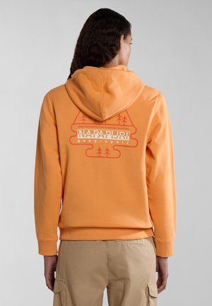 Personne aux longs cheveux foncés porte un sweat à capuche orange avec un graphisme Napapijri et un pantalon cargo beige, vue de dos sur fond gris.