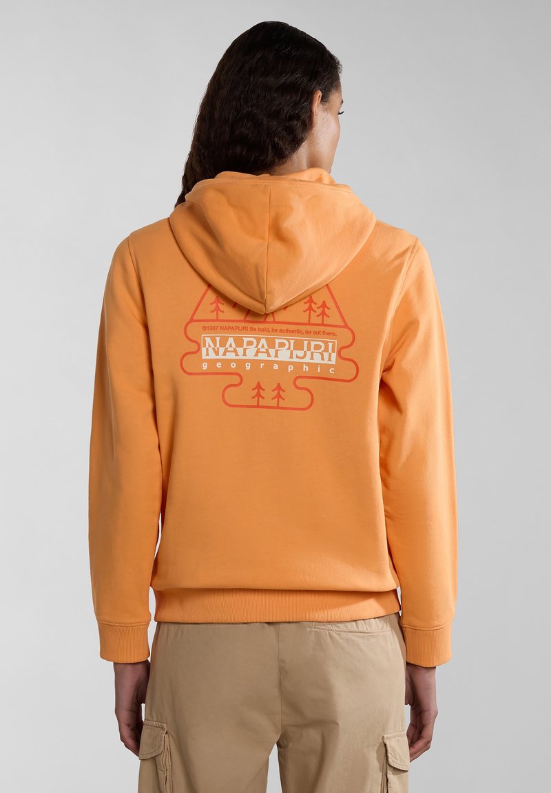 Personne aux longs cheveux foncés porte un sweat à capuche orange avec un graphisme Napapijri et un pantalon cargo beige, vue de dos sur fond gris.