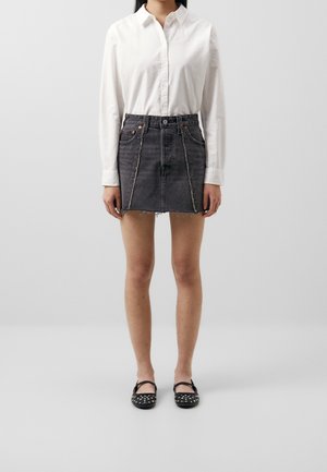 Personne portant une chemise blanche à manches longues boutonnée, une mini-jupe en denim noire avec des bords effilochés et des chaussures Mary Jane noires cloutées.