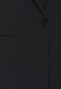 Fekete kabát egyetlen bekapcsolt fekete gombbal, bal oldalon zsebfedővel, sima, tiszta anyagtextúrával.