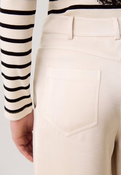 Pantalon beige avec poche arrière visible porté par une personne portant un haut à manches longues beige avec des rayures horizontales noires, main détendue le long du corps.