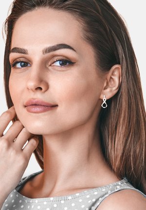 Silberne Unendlichkeitsohrringe an einer Person in einem grauen gepunkteten Oberteil, mit glattem Haar, neutralem Makeup und einer subtilen Hautstruktur.