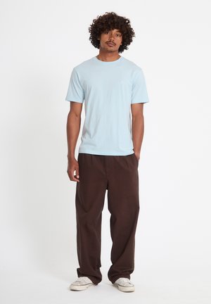 Nuori mies kiharatukkainen, yllään vaaleansininen lyhythihainen paita, väljät tummanruskeat housut ja valkoiset lenkkarit, seisoo yksinkertaisen taustan edessä.