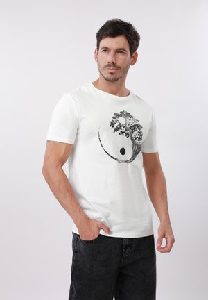 Rövid sötét hajú férfi fehér pólóban, amelyen fekete fa és absztrakt kör alakú minta látható, egyszerű háttér előtt pózol.