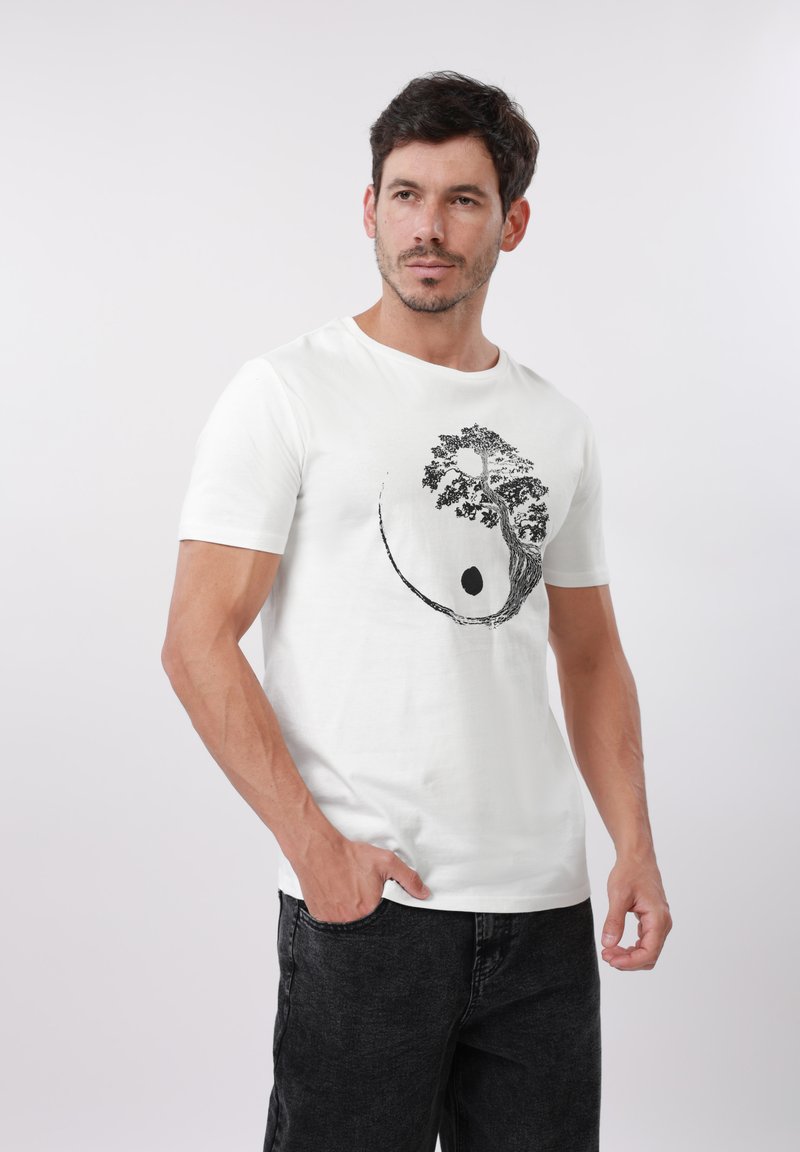 Άνδρας με κοντά σκούρα μαλλιά, που φοράει λευκό μπλουζάκι με μαύρο δέντρο και αφηρημένο κυκλικό σχέδιο, ποζάροντας μπροστά σε απλό φόντο.