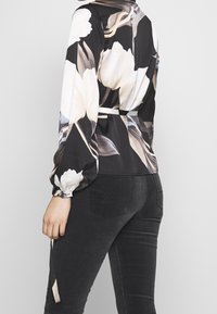 Zwarte blouse met grote bloemenprint, witte en beige bloemen, lange mouwen met elastische manchetten en een tailleband detail. Gepaarde met zwarte corduroy broek.