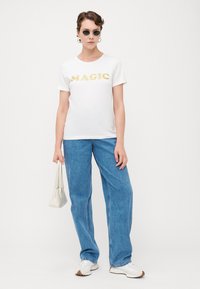 Hvit t-skjorte med "MAGIC" i gullglitter, kombinert med løse blå denimjeans og hvite sneakers. Holder en liten hvit håndveske.