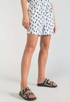 Pantalones cortos blancos con patrón ikat azul, cinturilla elástica y bolsillos laterales. Sandalias negras con tachuelas plateadas decorativas y correas.