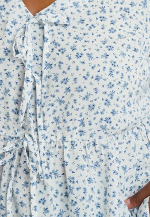 Close-up of white fabric with small blue floral print and tied bows worn by person with dark skin tone.