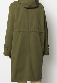 Olivgrön lång kappa med huva, midjehöjd som smalnar av, tryckknappsmanschetter och en slät yta. Bakvy, minimalistisk design.