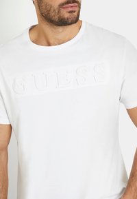 Tricou alb din bumbac cu un logo "GUESS" reliefat cu o textură fină pe față. Guler rotund și mâneci scurte.