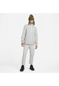 Γκρι φλις πουλόβερ με μισό φερμουάρ, ριχτές μανσέτες, λογότυπο στο στήθος; συνδυασμένο με ανοιχτό γκρι παντελόνι και μαύρα αθλητικά παπούτσια, με πράσινο σκουφί.