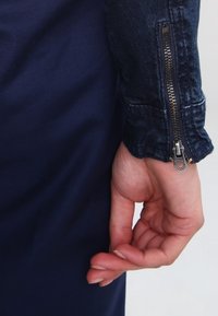 Manga de chaqueta de mezclilla oscura con un detalle de cremallera plateada. La tela tiene una textura ligeramente desgastada, y una mano descansa cerca del puño de la chaqueta.