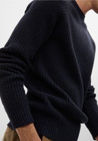 Homme portant un pull en maille texturée bleu marine foncé avec des poignets et un ourlet côtelés, associé à un pantalon marron clair.
